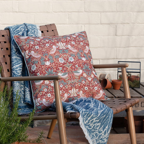 Strawberry Thief Floral Polyester Cushion in Berry on a wooden outdoor chair, deep red ground with mirrored birds, strawberries and foliage, 50x50cm, blue throw and terracotta pots nearby.