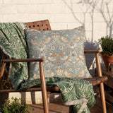 Strawberry Thief Floral Polyester Cushion in Biscuit, a square piped-edge cushion with grey-toned bird and berry print, shown on an outdoor wooden chair with a green patterned throw.