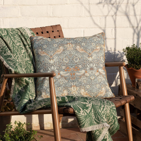 Strawberry Thief Floral Polyester Cushion in Biscuit, a square piped-edge cushion with grey-toned bird and berry print, shown on an outdoor wooden chair with a green patterned throw.