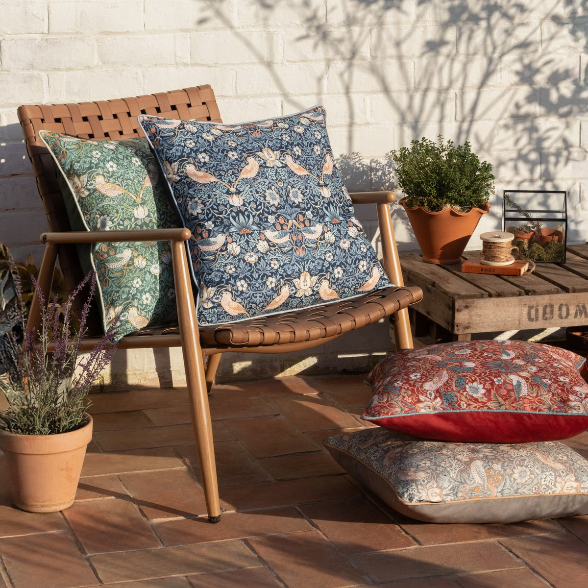 Strawberry Thief Floral Polyester Cushion in Biscuit, blue-toned square design with piped edge, styled on an outdoor patio chair alongside matching green and red patterned cushions.