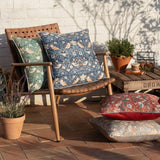 Strawberry Thief Floral Polyester Cushion in Biscuit, blue-toned square design with piped edge, styled on an outdoor patio chair alongside matching green and red patterned cushions.