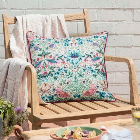 Strawberry Thief Floral Polyester Cushion in coral and leaf green, printed with birds, strawberries and foliage, coral piped edge, shown on a wooden garden bench.
