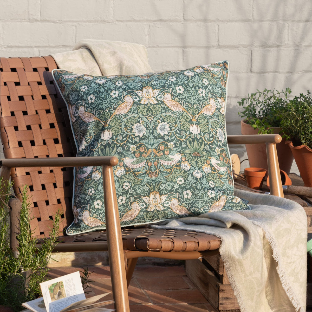 Strawberry Thief Floral Polyester Cushion in Forest Green, 50 x 50cm square with piped edge trim, styled on a wooden outdoor chair with cream throws and potted plants.