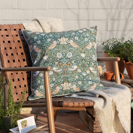 Strawberry Thief Floral Polyester Cushion in Forest Green, 50 x 50cm square with piped edge trim, styled on a wooden outdoor chair with cream throws and potted plants.