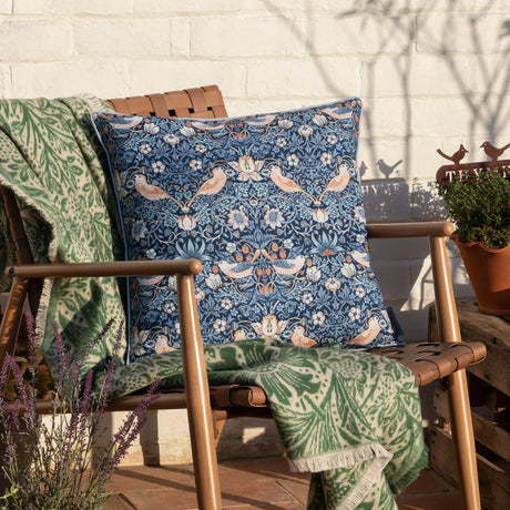 Strawberry Thief Floral Polyester Cushion in indigo with bird and floral print, pale piped trim, styled on an outdoor wooden chair with a green throw.