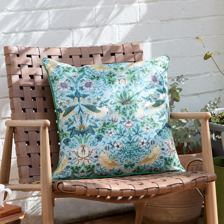 Strawberry Thief Floral Polyester Cushion in Mineral/Yellow, square with bird and floral print, shown on a woven leather chair against a white brick wall.