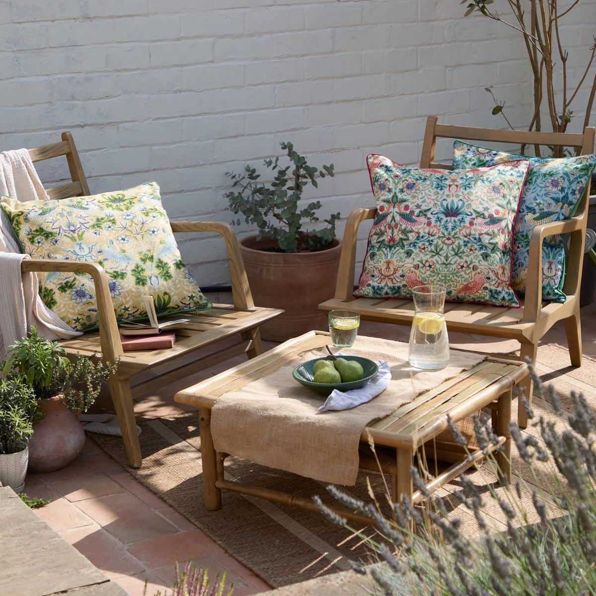 Strawberry Thief Floral Polyester Cushions in mineral-blue and yellow with bird and floral patterns, styled on wooden garden chairs alongside a coffee table outdoors.