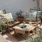 Strawberry Thief Floral Polyester Cushions in mineral-blue and yellow with bird and floral patterns, styled on wooden garden chairs alongside a coffee table outdoors.