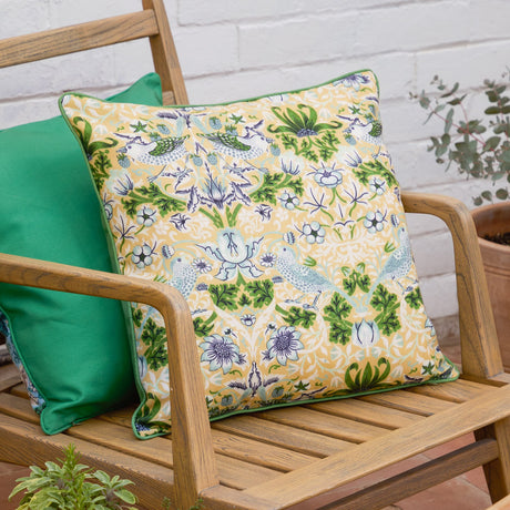 Strawberry Thief Floral Polyester Cushion in Sunflower, square with green piping and bird and floral pattern on yellow, shown on a wooden garden bench with a plain green cushion.