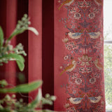 Berry red velvet pencil pleat curtain shown pleated near a window, with an embroidered Strawberry Thief leading edge featuring birds, strawberries and foliage.
