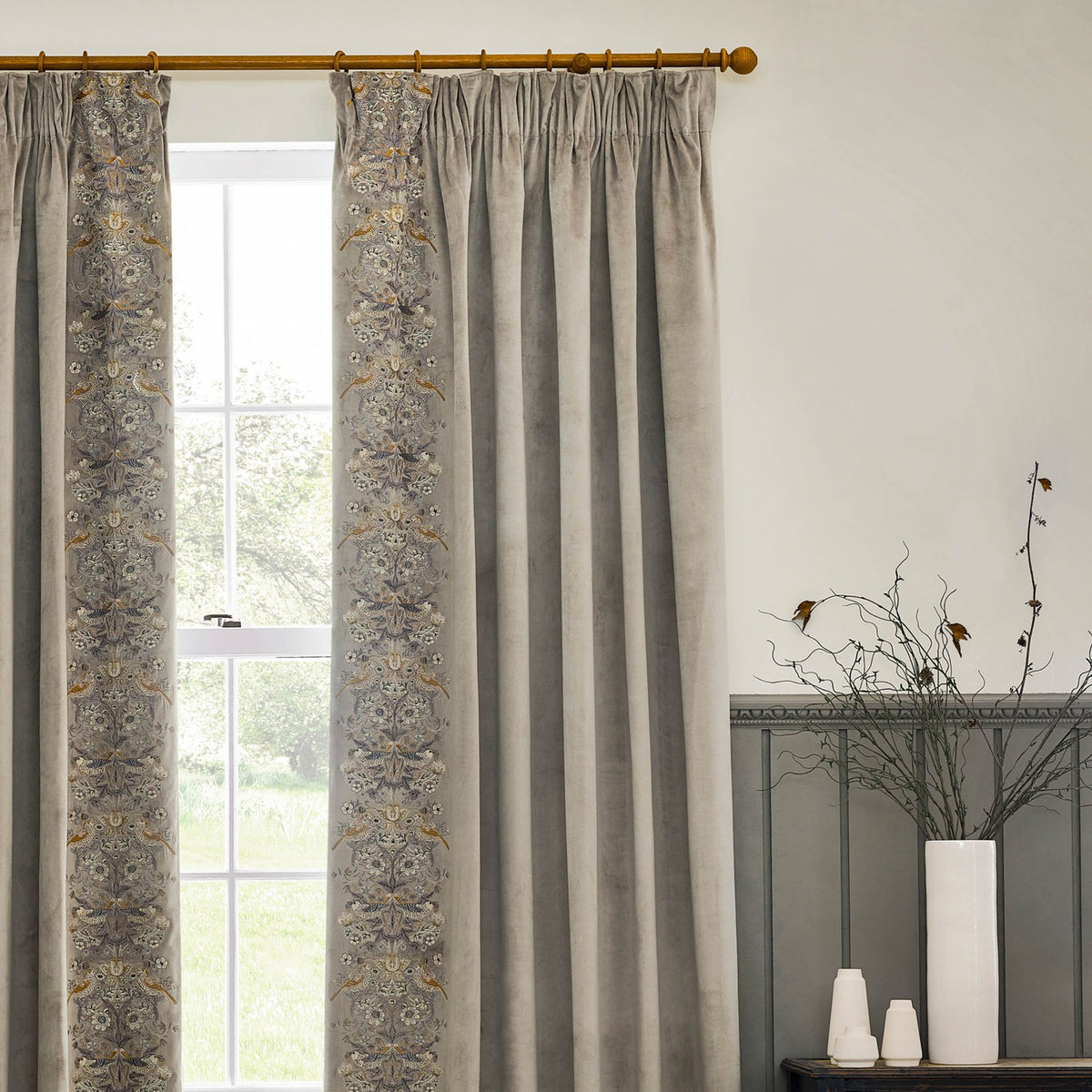 Strawberry Thief Velvet Pencil Pleat Curtains in biscuit, hung at a window with pencil pleat heading and an embroidered leading edge featuring birds and floral motifs.