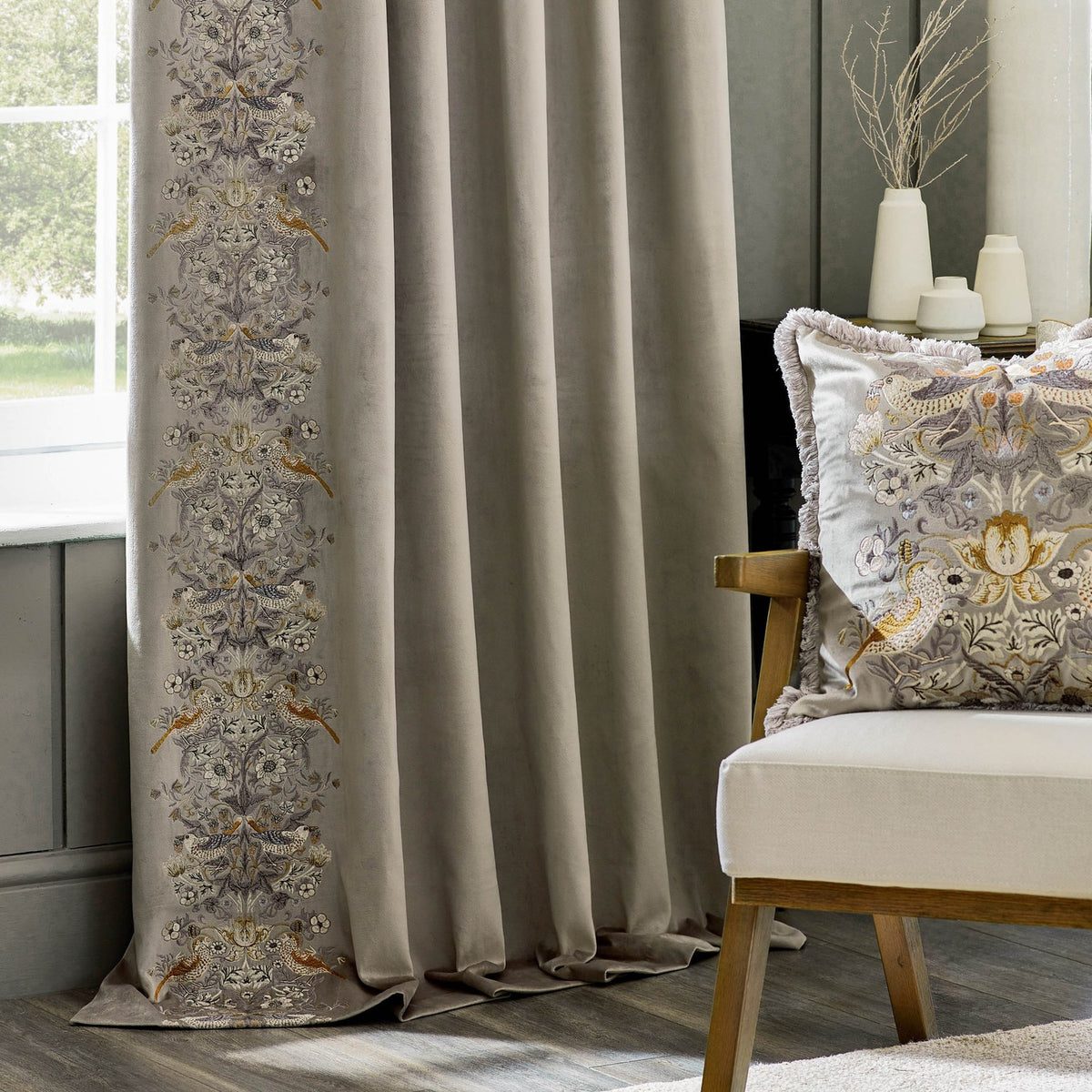 Biscuit velvet pencil pleat curtain with embroidered Strawberry Thief bird and floral leading edge, shown pencil pleated in a living room beside a cushioned armchair.
