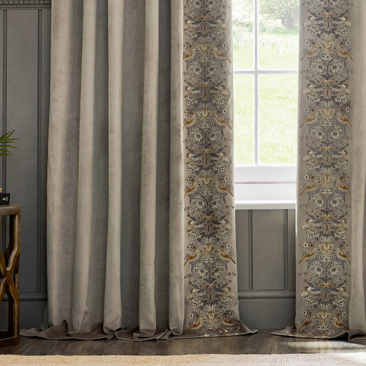Strawberry Thief pencil pleat curtains in biscuit velvet with embroidered bird and floral leading edge, shown pleated beside a window overlooking greenery.