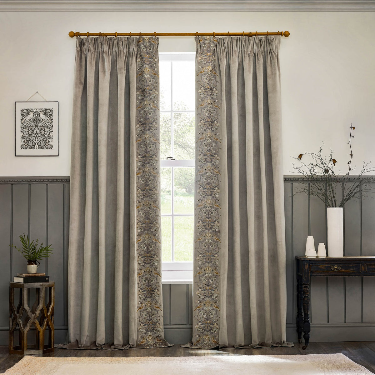 Strawberry Thief Velvet Pencil Pleat Curtains in biscuit, with embroidered floral and bird leading edge, hung on a gold pole in a living room with panelled walls.