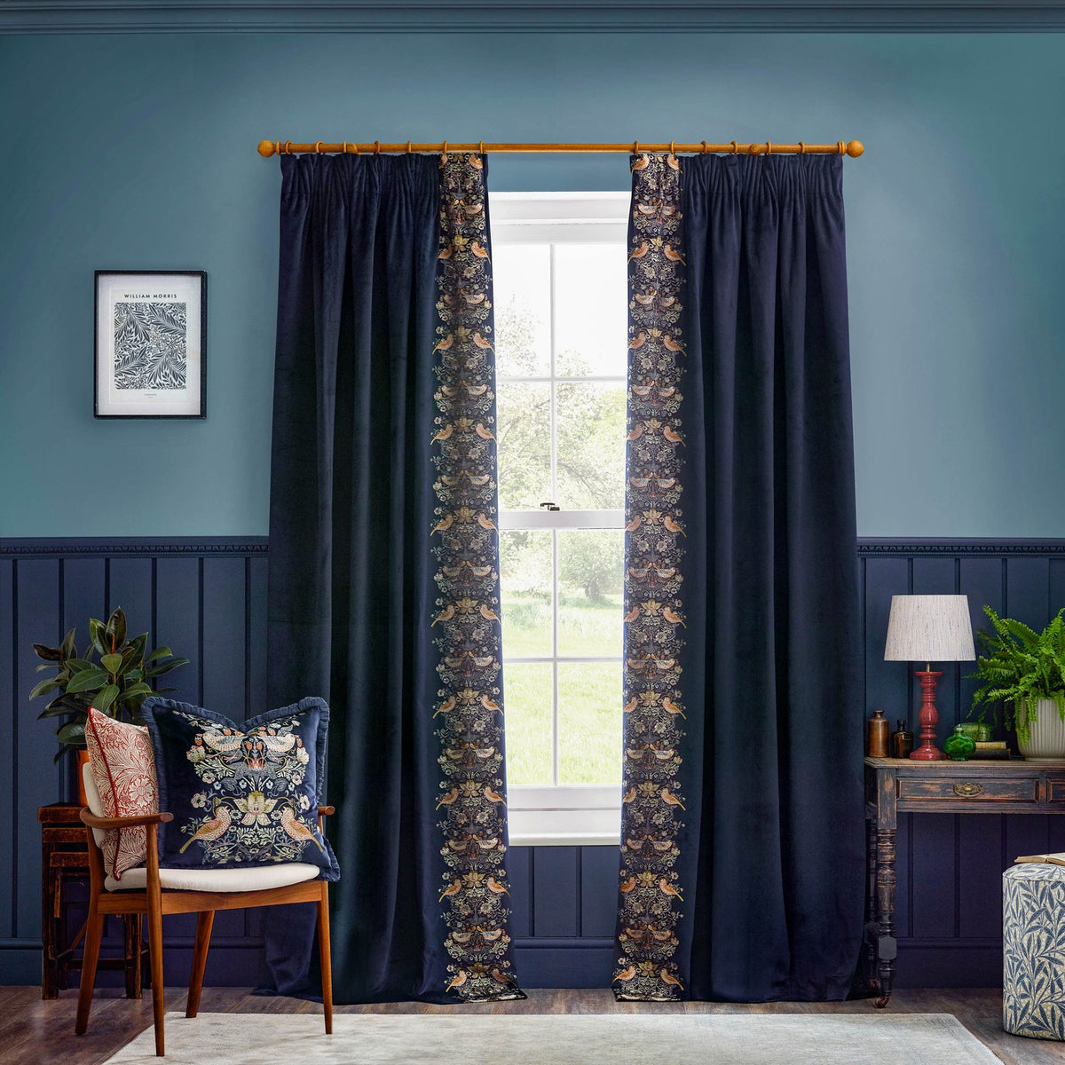 Indigo velvet pencil pleat curtains with Strawberry Thief bird and floral embroidery along the leading edge, styled either side of a window in a blue-toned living room.