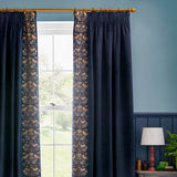 Indigo velvet pencil pleat curtains with a Strawberry Thief embroidered bird and floral border, hung on a wooden pole in a blue-walled living room.
