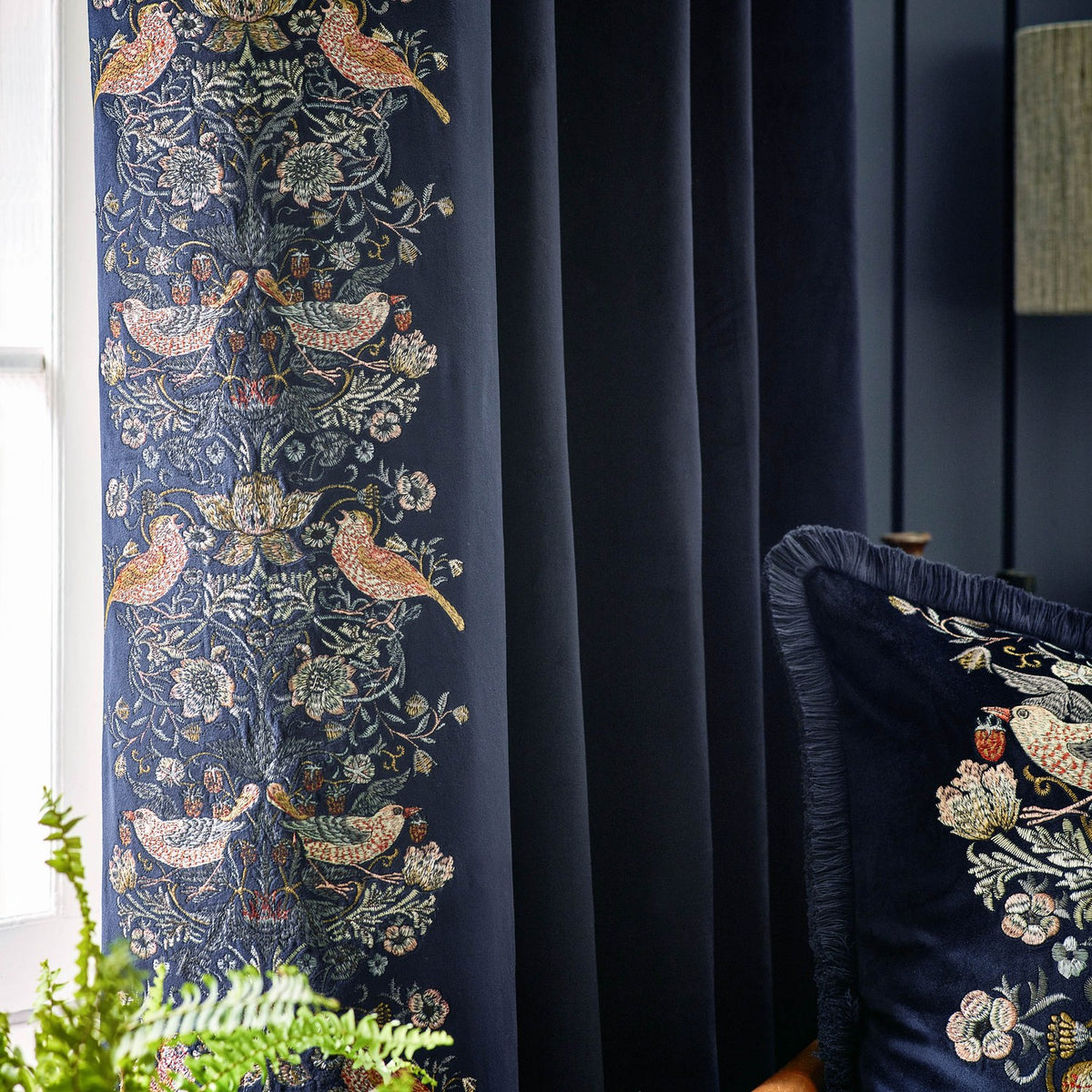 Indigo velvet pencil pleat curtain with Strawberry Thief embroidered bird and floral border, shown pleated by a window beside a matching fringed cushion.