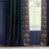 Indigo velvet pencil pleat curtains with an embroidered Strawberry Thief bird and floral border in rust, gold and cream along the leading edge, shown hanging beside a window.