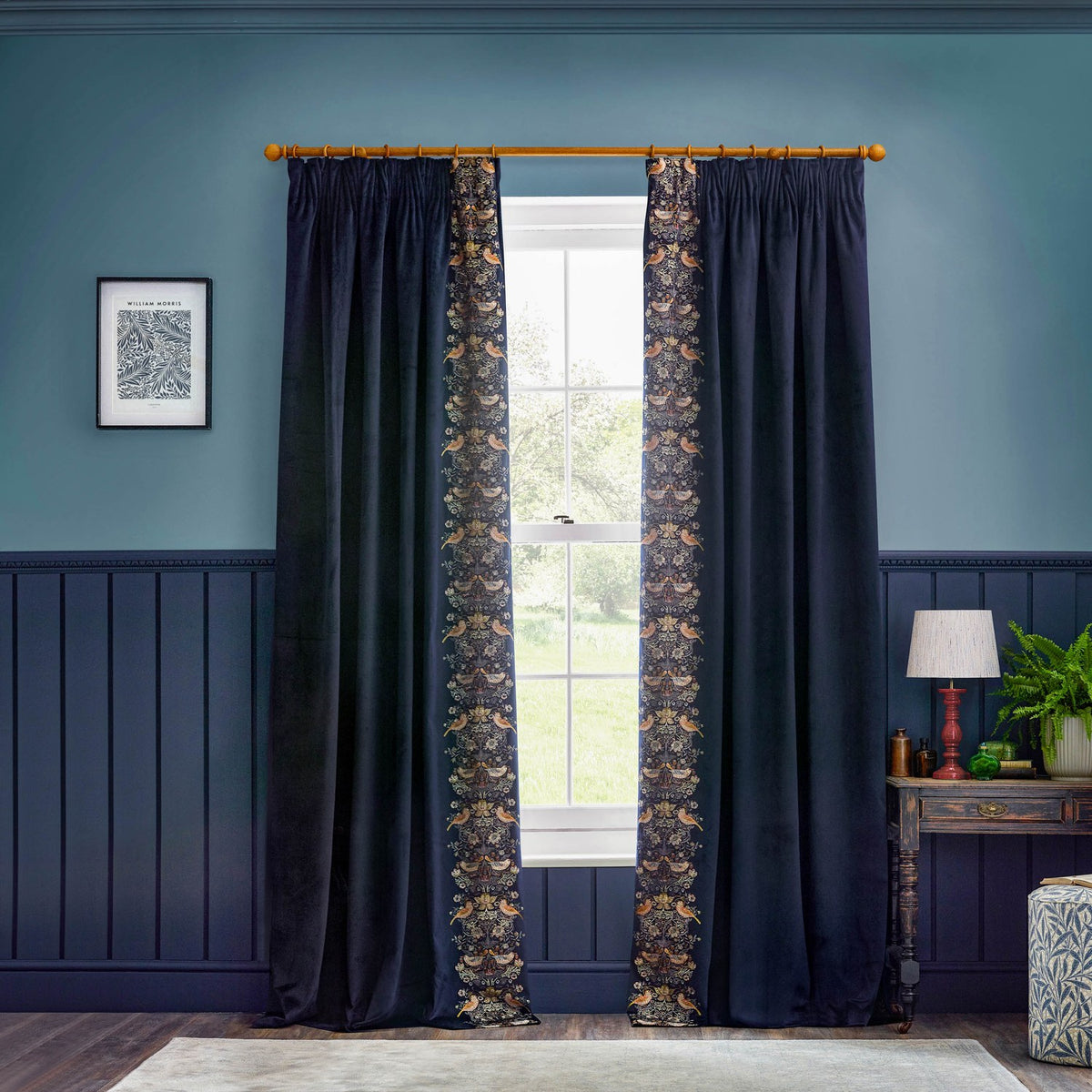 Indigo velvet pencil pleat curtains with Strawberry Thief embroidered leading edge in rust, gold and sage, hung on a wooden pole in a blue-panelled room.