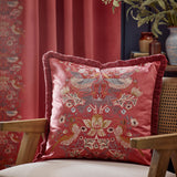 Strawberry Thief Jacquard Velvet Cushion in Berry, a square red velvet cushion with embroidered bird and floral pattern and fringed edges, shown on a chair in a living room setting.