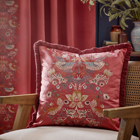 Strawberry Thief Jacquard Velvet Cushion in Berry, a square red velvet cushion with embroidered bird and floral pattern and fringed edges, shown on a chair in a living room setting.