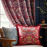 Strawberry Thief Jacquard Velvet Cushion in berry red with fringed trim, featuring bird and floral embroidery, styled on an armchair beside patterned curtains.