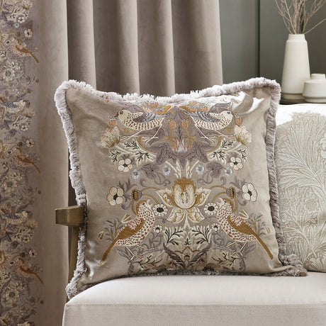 Strawberry Thief Jacquard Velvet Cushion in biscuit-beige, with bird and floral embroidery in gold and grey, fringed edges, shown on a chair beside curtains in a matching print.