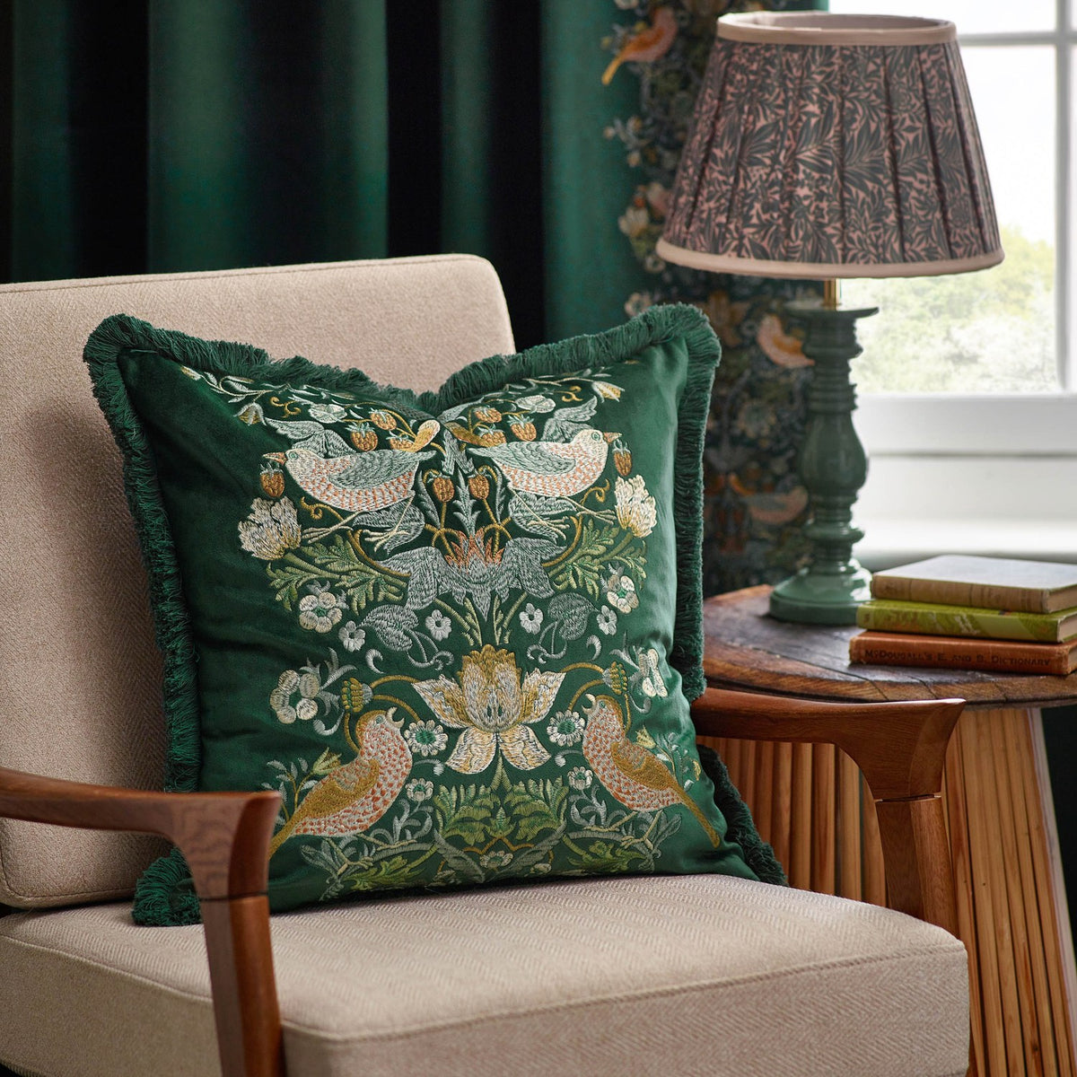 Strawberry Thief jacquard velvet cushion in forest green with embroidered bird and floral design, fringe trim, styled on a beige armchair beside a lamp.
