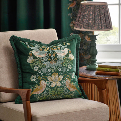 Strawberry Thief jacquard velvet cushion in forest green with embroidered bird and floral design, fringe trim, styled on a beige armchair beside a lamp.