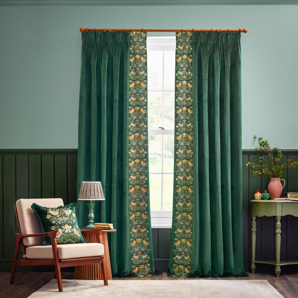 Strawberry Thief Jacquard Velvet Cushion in Forest Green on a beige armchair, featuring embroidered birds, florals and strawberries in cream, gold and rust in a living room setting.