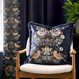 Strawberry Thief Jacquard Velvet Cushion in indigo, fringed square cushion embroidered with mirrored bird and strawberry motifs, shown on an armchair against dark curtains.