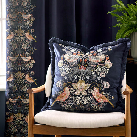 Strawberry Thief Jacquard Velvet Cushion in indigo, fringed square cushion embroidered with mirrored bird and strawberry motifs, shown on an armchair against dark curtains.