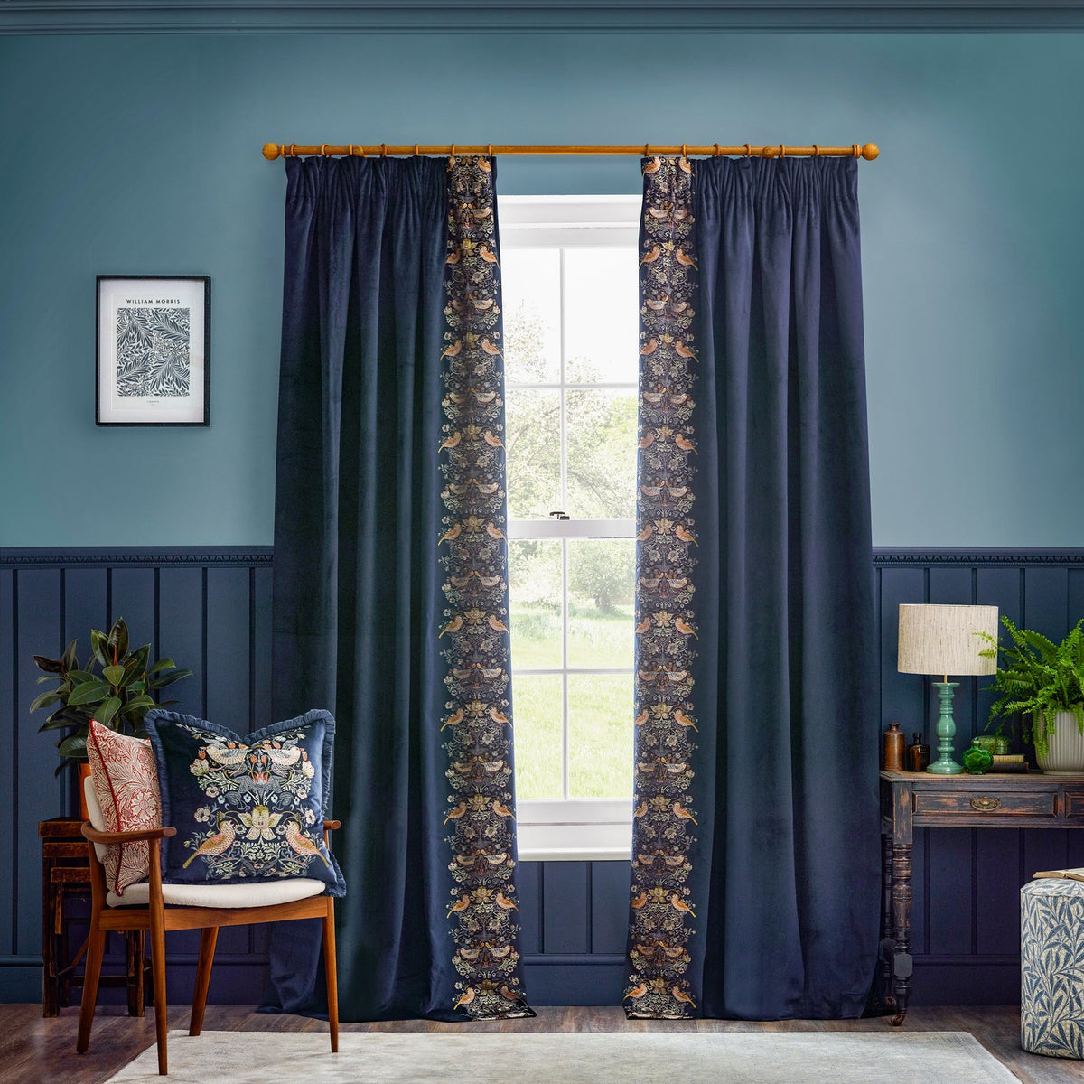 Strawberry Thief Jacquard Velvet Cushion in indigo on a wooden armchair, showing mirrored bird and floral embroidery in cream, gold and rust on a deep blue velvet ground.