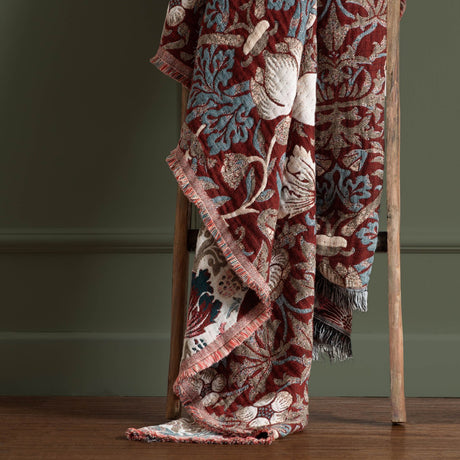 Strawberry Thief Jacquard Cotton Throw in berry red draped over a wooden ladder, showing bird and floral pattern in blue and cream with fringed edge.