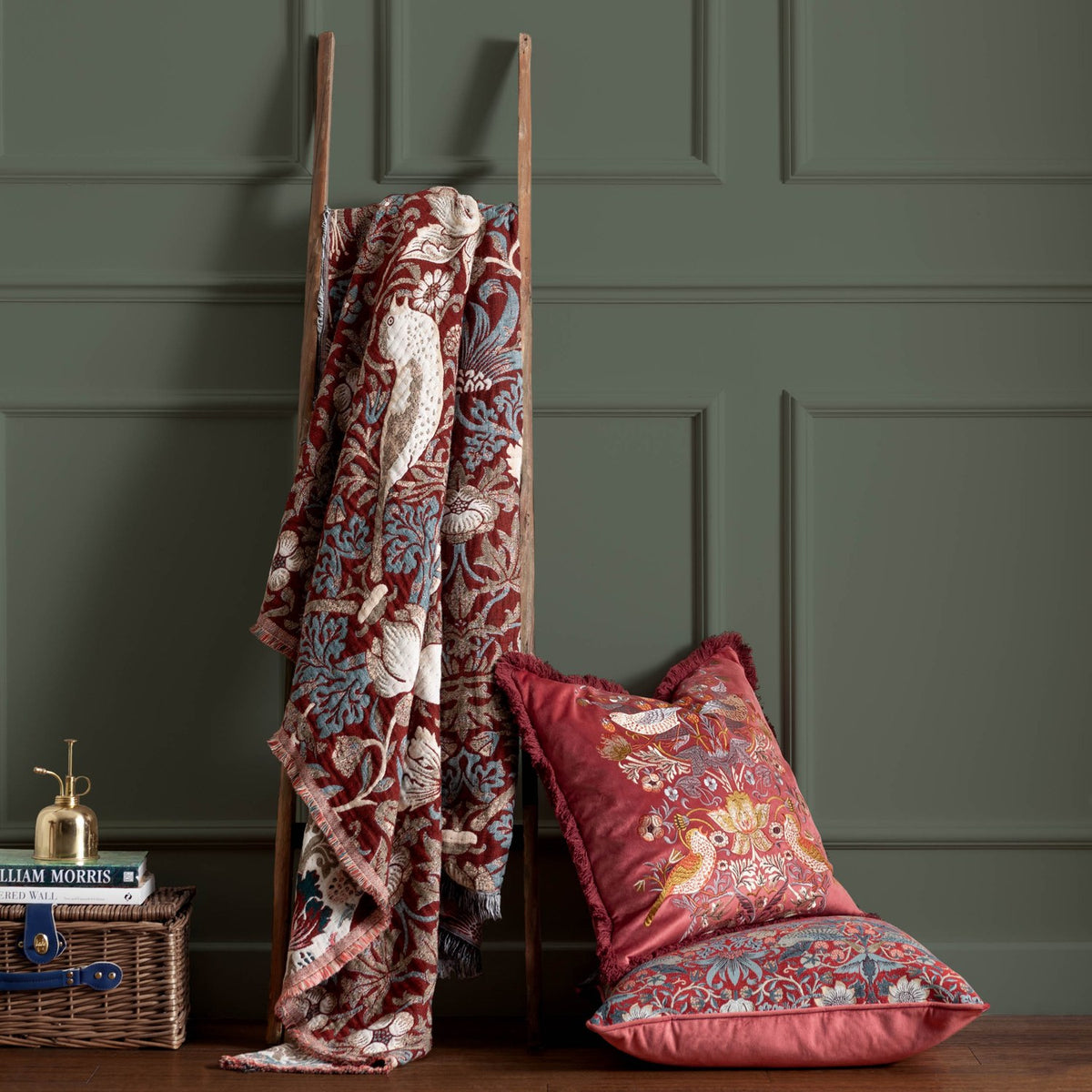 Strawberry Thief Jacquard Cotton Throw in berry-red, draped over a wooden ladder, showing bird and floral motifs in cream and blue, styled beside matching cushions.