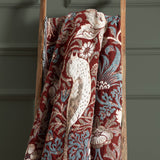 Strawberry Thief Jacquard Cotton Throw in Berry, draped over a wooden ladder, showing bird and floral motifs in cream and blue on a deep red ground.