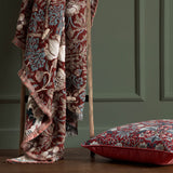 Strawberry Thief Jacquard Cotton Throw in Berry draped over a wooden ladder, showing bird and floral pattern in red, blue and cream with fringed edge, beside a matching cushion on a wooden floor.
