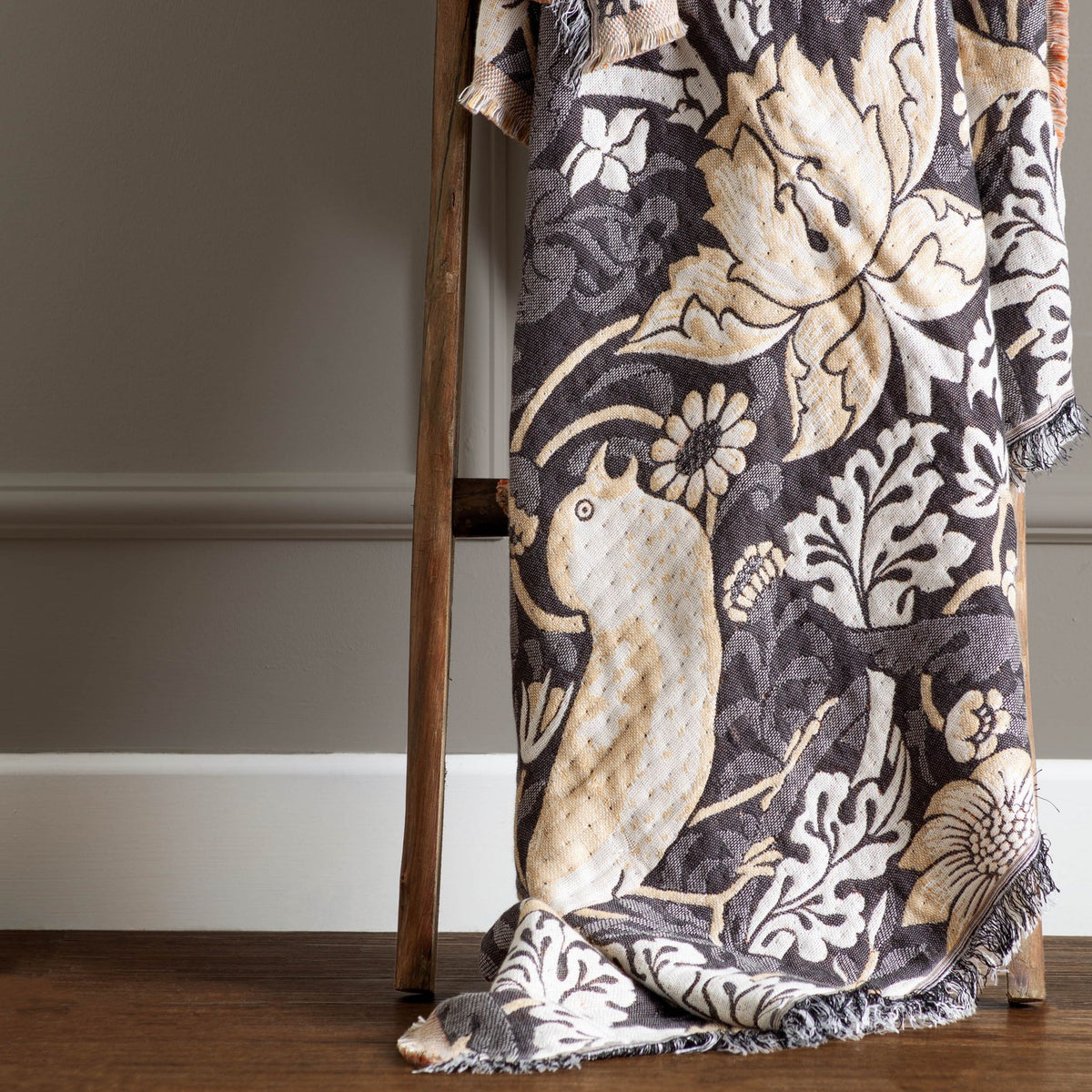 Strawberry Thief Jacquard Cotton Throw in biscuit, charcoal ground with cream bird and floral motifs, fringed edge, draped over a wooden ladder.