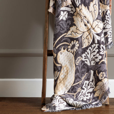 Strawberry Thief Jacquard Cotton Throw in biscuit, charcoal ground with cream bird and floral motifs, fringed edge, draped over a wooden ladder.