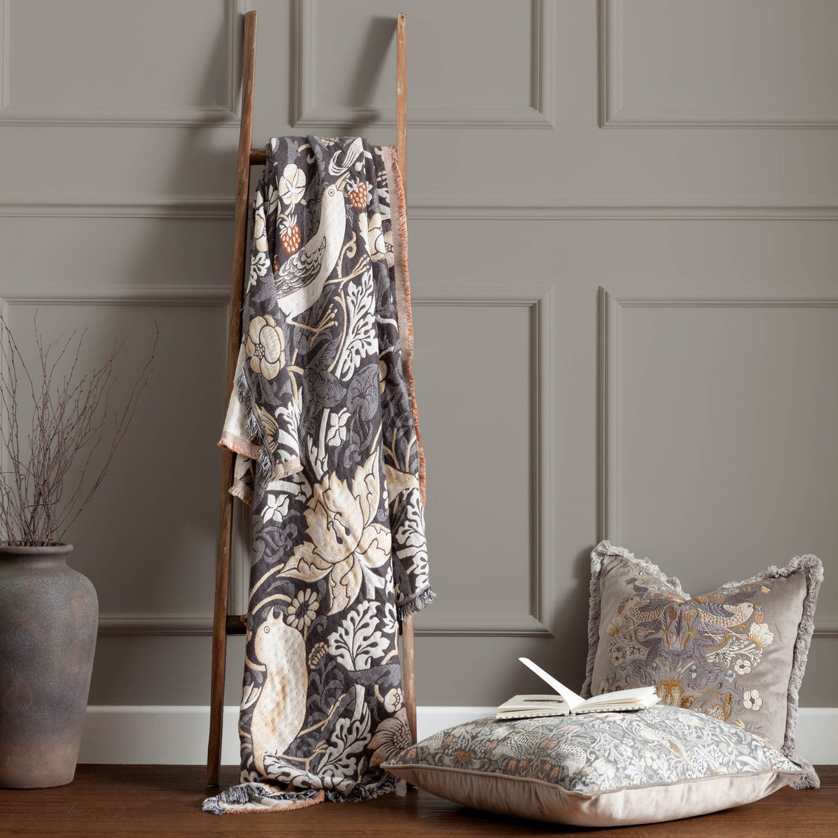 Strawberry Thief Jacquard Cotton Throw in Biscuit draped over a wooden ladder, showing a charcoal ground with bird, floral and strawberry motifs, styled beside cushions in a living room setting.