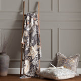Strawberry Thief Jacquard Cotton Throw in Biscuit draped over a wooden ladder, showing a charcoal ground with bird, floral and strawberry motifs, styled beside cushions in a living room setting.