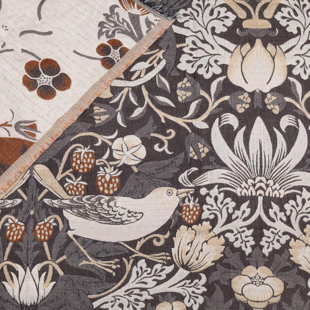 Strawberry Thief Jacquard Cotton Throw in biscuit, showing the folded corner reverse in cream with rust florals against the charcoal jacquard weave of birds, strawberries and flowers.
