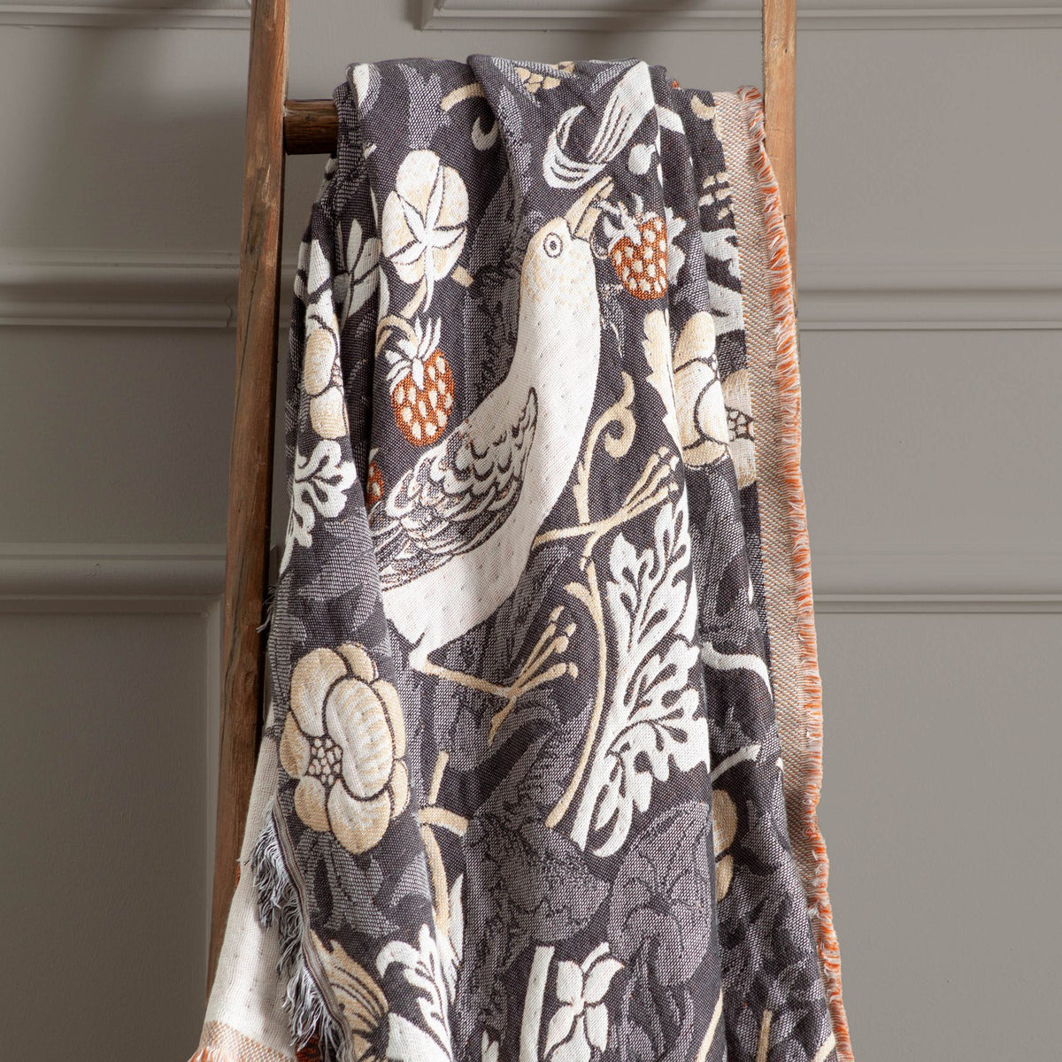 Strawberry Thief Jacquard Cotton Throw in Biscuit draped over a wooden ladder, showing a charcoal ground woven with birds, flowers and strawberries, with contrasting fringed edge.