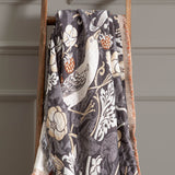 Strawberry Thief Jacquard Cotton Throw in Biscuit draped over a wooden ladder, showing a charcoal ground woven with birds, flowers and strawberries, with contrasting fringed edge.
