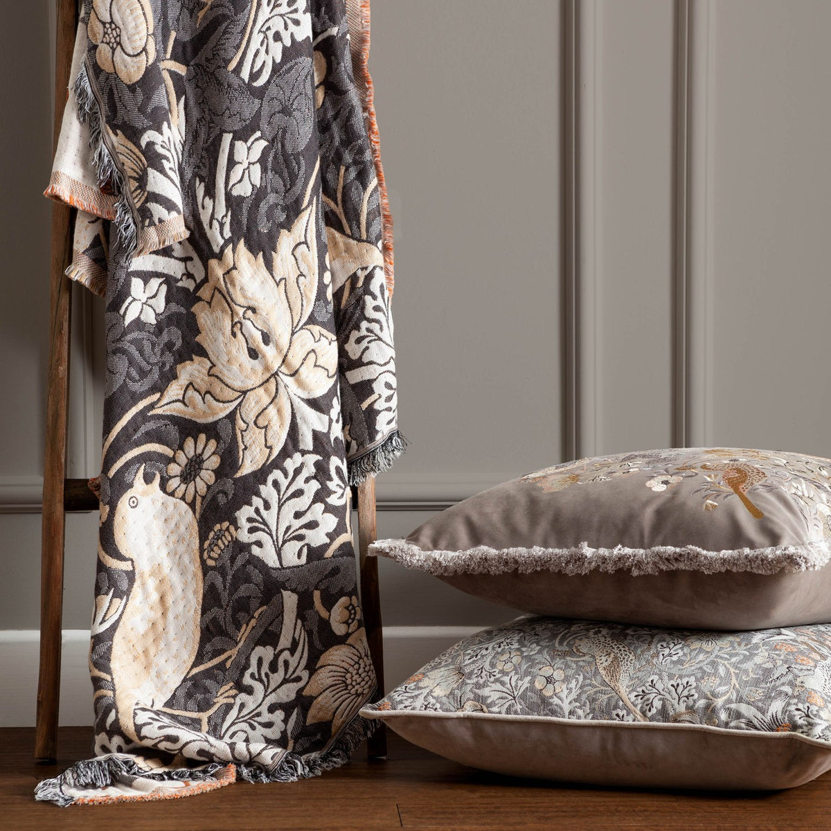 Strawberry Thief Jacquard Cotton Throw in Biscuit, charcoal ground with bird and floral motifs and fringed edge, draped over a wooden ladder beside two patterned cushions.