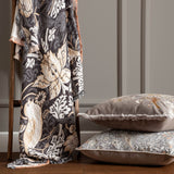 Strawberry Thief Jacquard Cotton Throw in Biscuit, charcoal ground with bird and floral motifs and fringed edge, draped over a wooden ladder beside two patterned cushions.