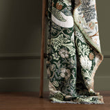 Strawberry Thief Jacquard Cotton Throw in Forest Green draped over a wooden ladder, showing bird and floral pattern with cream, green and rust tones and fringed edge.