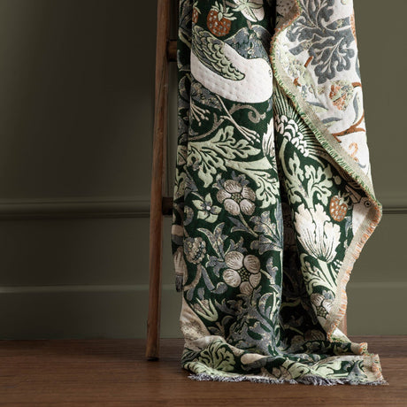 Strawberry Thief Jacquard Cotton Throw in Forest Green draped over a wooden ladder, showing bird and floral pattern with cream, green and rust tones and fringed edge.