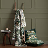 Strawberry Thief Jacquard Cotton Throw in forest green, draped over a wooden ladder with bird and floral pattern, styled beside matching cushions and a side table.