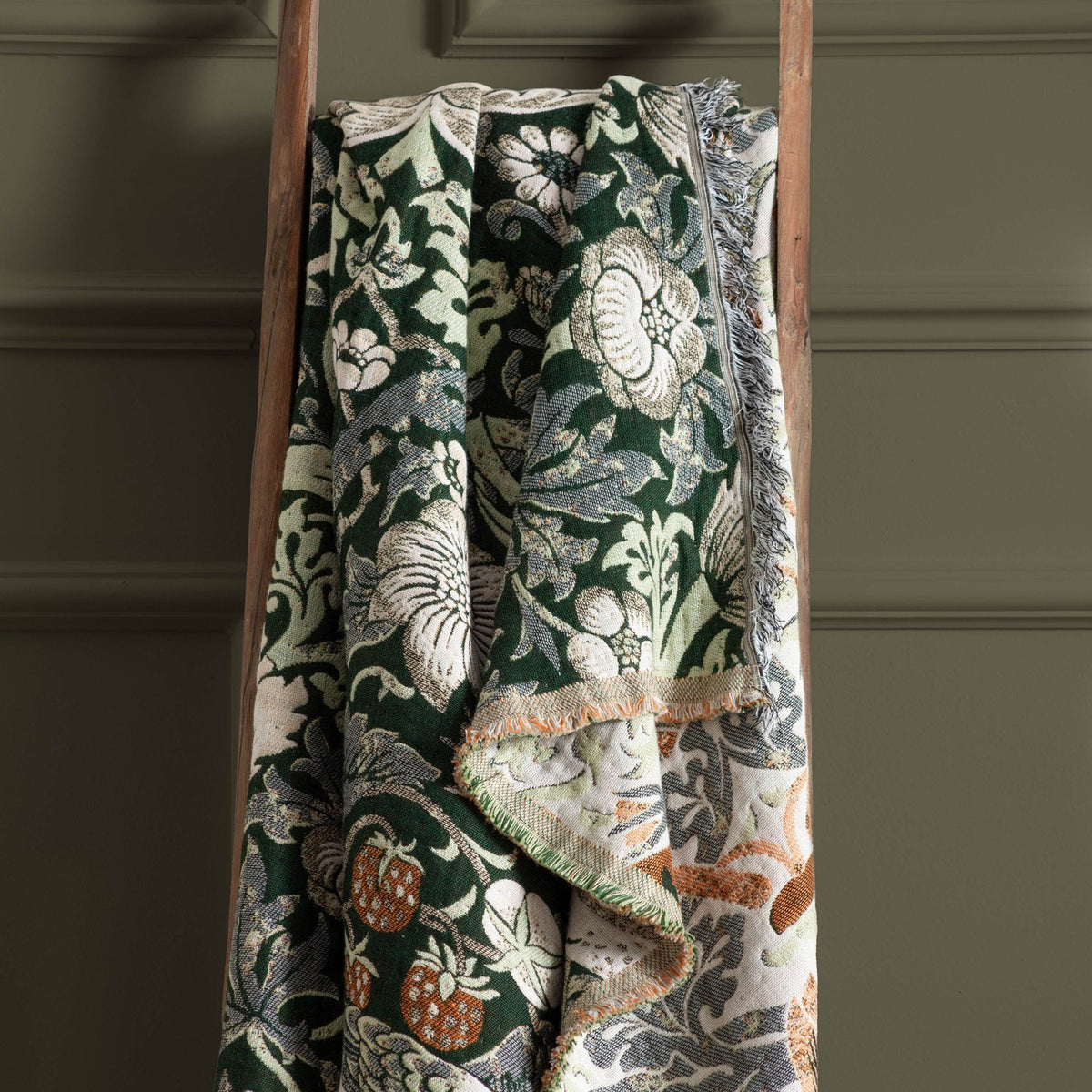 Strawberry Thief Jacquard Cotton Throw in Forest Green, draped over a wooden ladder, showing the William Morris bird, strawberry and floral pattern with fringed edges.